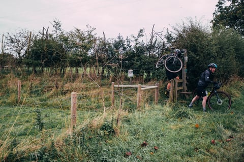 A Gravel Bonanza rider lifts a gravel bike over a wooden stile while another pushes through the gate in the Peak District.