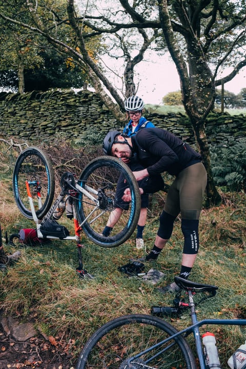 Gravel Bonanza rider repairs a bike turned upside down on the grass by a drystone wall while another waits.