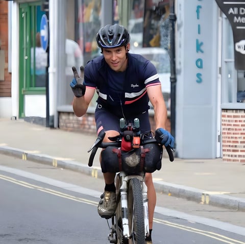 Chris Hinds rides a loaded bike through a town and flashes a peace sign during the Great British Divide.