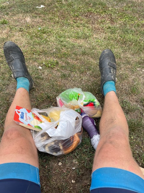 Rider's legs and dusty cycling shoes stretched out on grass beside bags of Hungarian shop food, cheese and pastries.