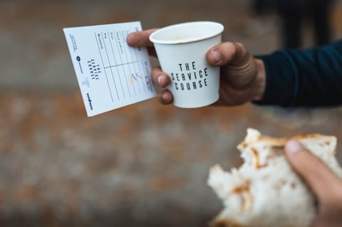 Hand holds a Service Course paper cup and a Gravel Bonanza ride card with boxes for coffee, feed station, pie and pint.