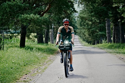Hungarian Divide rider on a bikepacking bike with a handlebar bag rides a tree-lined tarmac lane in summer.