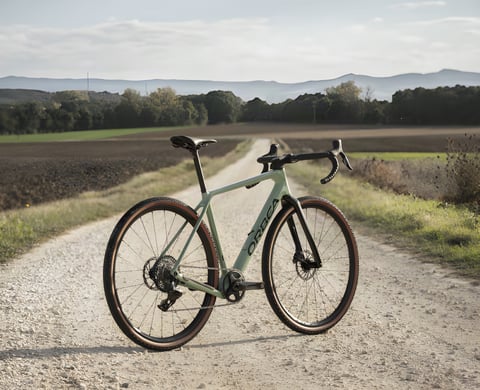 2022 Orbea Terra gravel bike in sage green parked on a white gravel track between fields, hills behind.