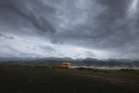 Yellow minibus raises dust on a track beneath storm clouds and mountains in Kyrgyzstan.