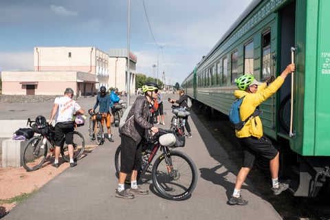 Silk Road Mountain Race riders lift loaded bikes onto a green train at a station in Kyrgyzstan.