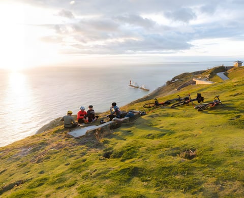 Glampacking riders rest on a grassy Isle of Wight clifftop at sunset, bikes on the grass and the Needles offshore.