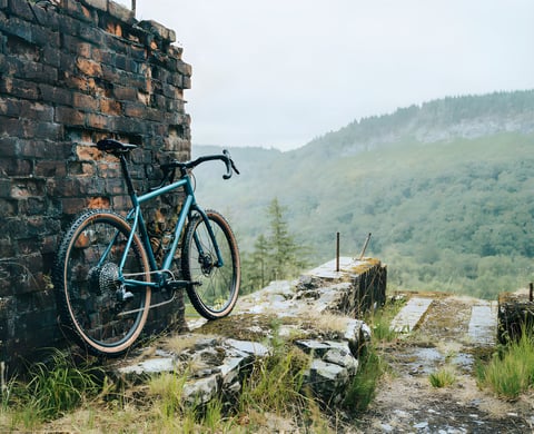 Ribble Gravel 725 steel gravel bike leans against a brick ruin, with wooded hills behind.