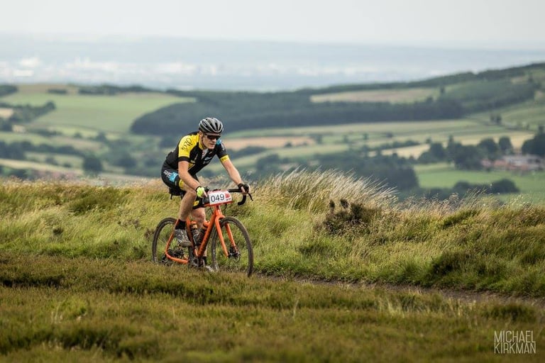 James on his orange Mason Bokeh, race number 049, climbs a grassy moorland trail at Yorkshire True Grit.
