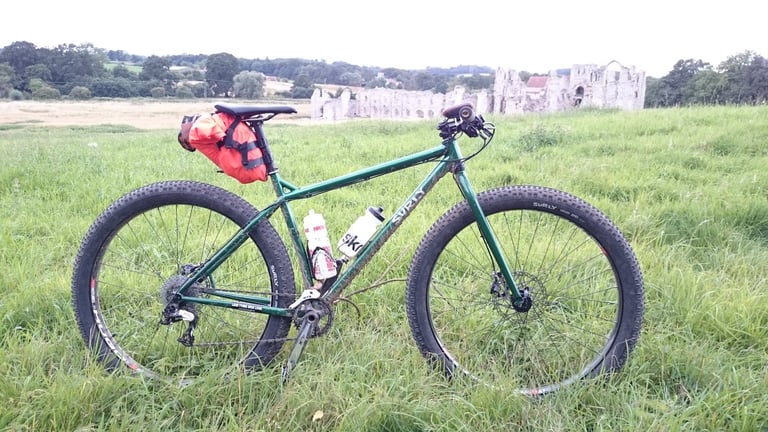 Green Surly Krampus with an orange seatpack stands in long grass in front of the ruins of Castle Acre Priory, Norfolk.