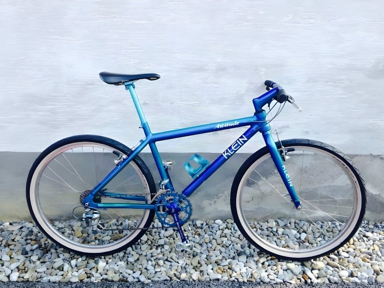 Blue Klein Attitude mountain bike leaning against a white wall above a bed of pebbles.