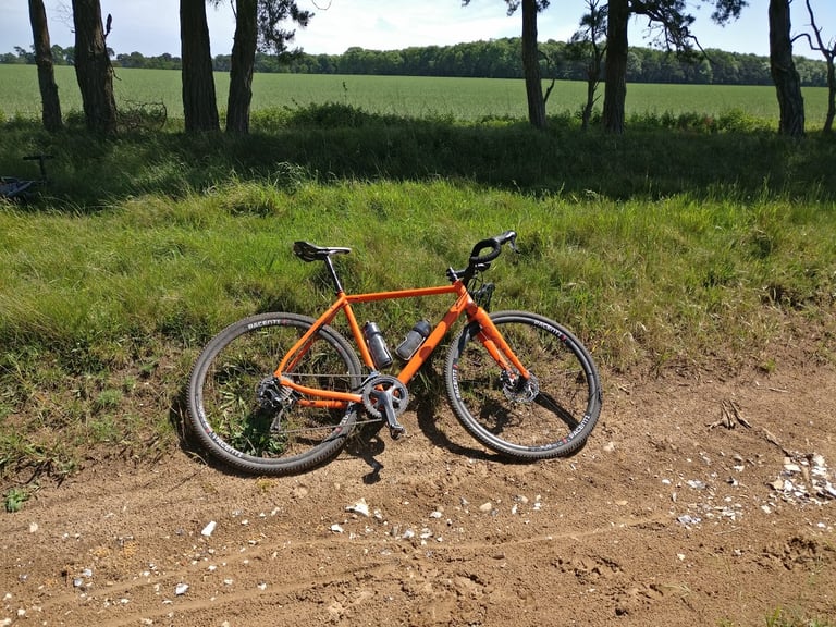 Orange gravel bike with drop bars and disc brakes lying on a grass verge beside a sandy farm track.