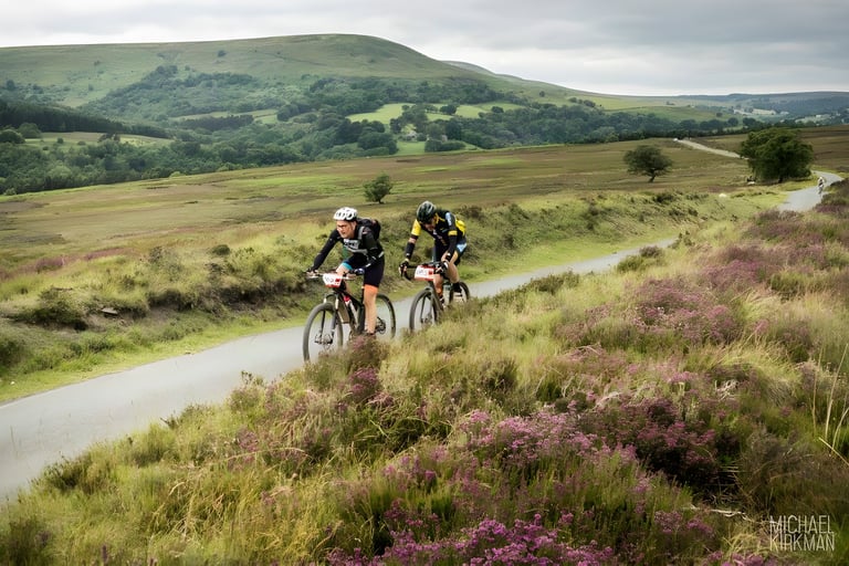 Two riders pedal a tarmac lane through flowering heather on the Yorkshire True Grit course, hills behind.