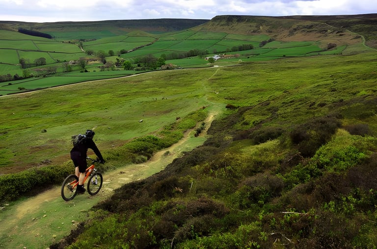 Rider with a rucksack on an orange mountain bike follows a narrow moorland trail above green farmland.