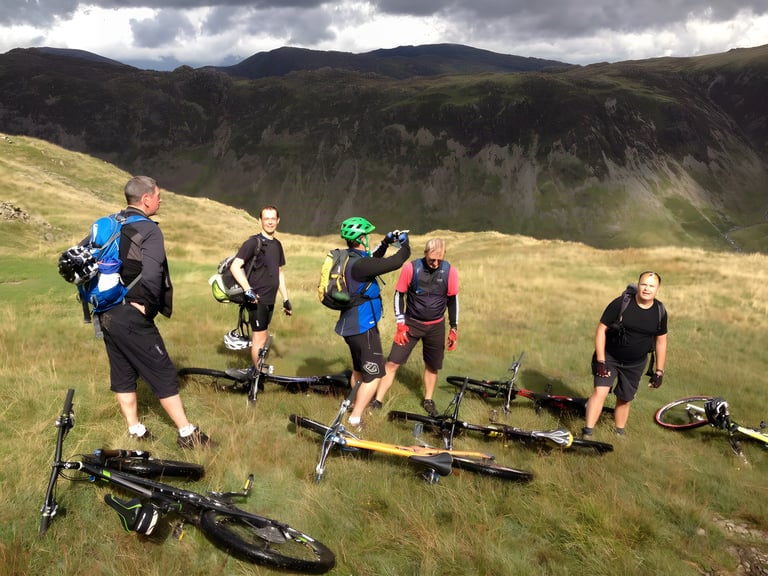 Chasing Trails group of mountain bikers rest on a grassy fellside, bikes laid down, craggy hills behind.