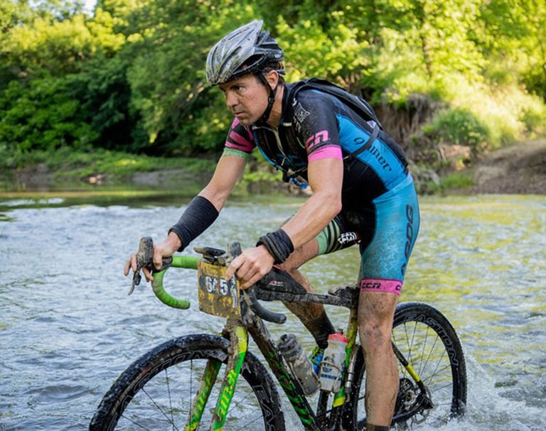 Mud-splattered racer rides a gravel bike with green-taped Easton EC70 AX bars through a shallow river.