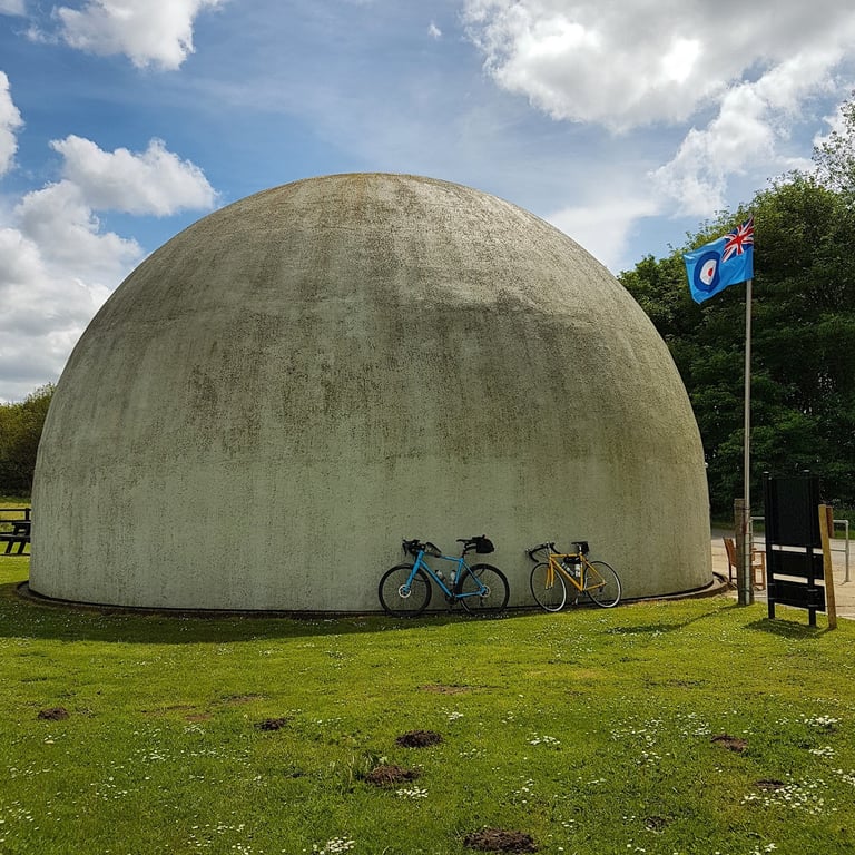 Two road bikes leaning against the concrete Langham Dome in Norfolk on an Audax ride, an RAF flag flying beside it.