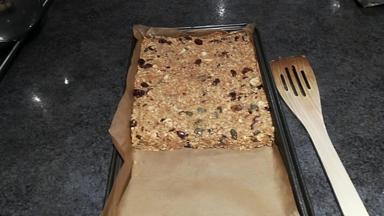 Homemade energy bar mixture pressed into half a lined baking tray, ready for the oven, with a wooden spatula.