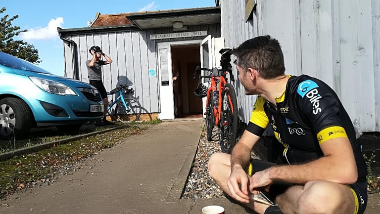 Rider crouching outside Blaxhall Village Hall at the Suffolk Byways Audax, with bikes leaning against the wall.