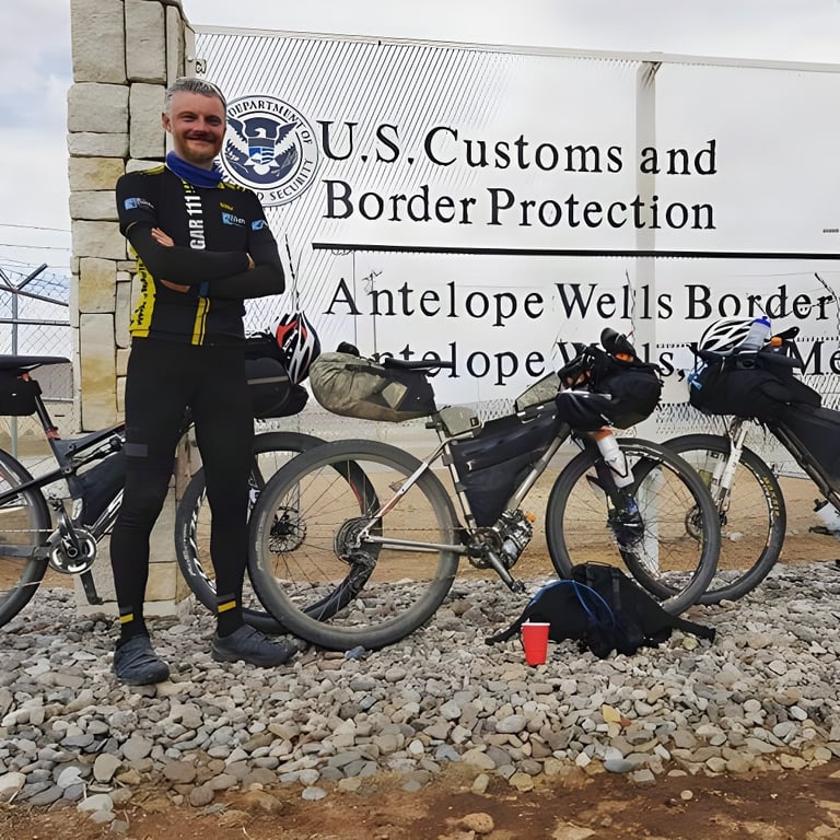 Rider finishes the Tour Divide at the US Customs and Border Protection sign at Antelope Wells, with loaded bikes behind.