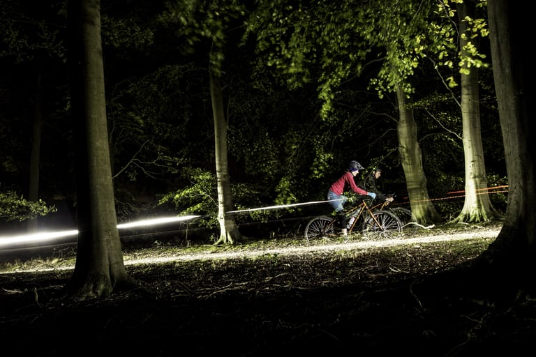 Long-exposure shot of two night riders in woodland, a streak of light trailing from their lights along the track.