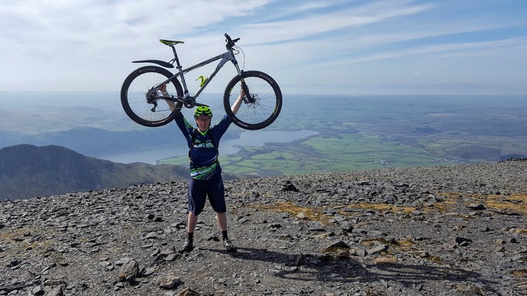 Bikepacking beginner James Pearson lifts his mountain bike above his head on a Lake District fell top.