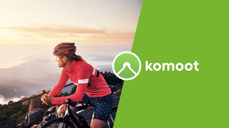 Komoot logo beside a cyclist in a red helmet and jersey resting on the bars above a hazy view of sea and hills.