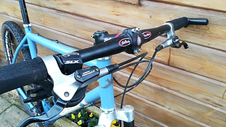 Singular Swift cockpit with a flat Salsa bar, Shimano SLX shifters and bar ends, against a wooden cabin wall.