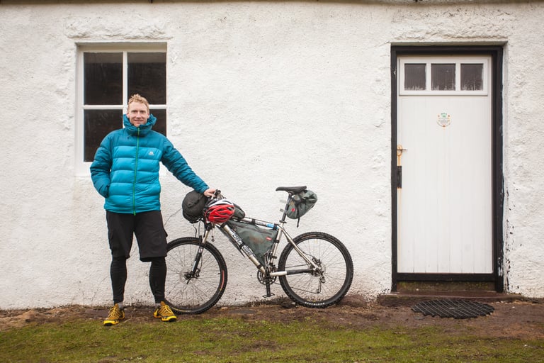 Alastair Humphreys beside his Orange bikepacking rig, fitted with frame and seat bags, against a bothy wall.