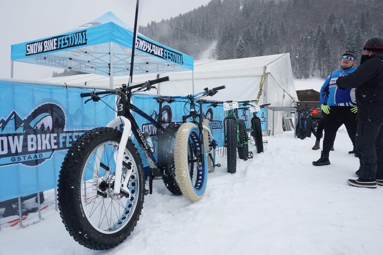 Fat bikes parked beside Snow Bike Festival Gstaad banners as riders chat in the snow before the prologue.