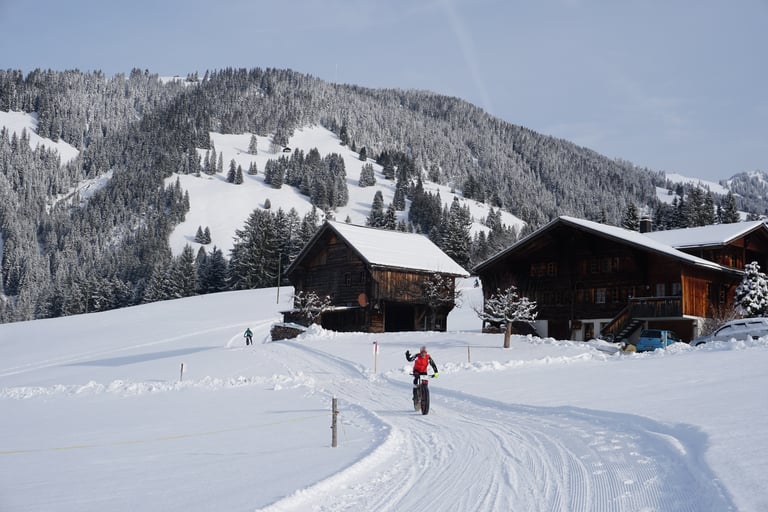 Fatbike rider descends a groomed snow track past timber chalets and wooded slopes in Switzerland, Snow Bike Festival.