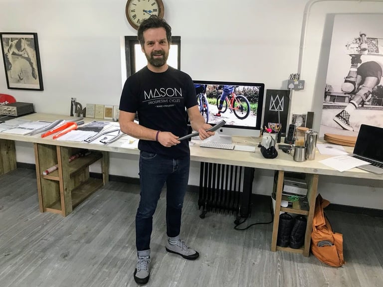 Dom Mason holds a length of frame tubing in the Mason Cycles office, bent tubes and a skateboarding print behind him.
