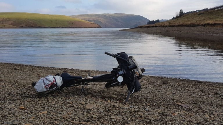 Loaded bike resting on its side by the water's edge at Clywedog Reservoir, with green hills across the lake.