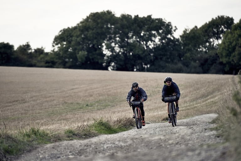 Two riders on Mason bikes with handlebar bags ride a gravel track beside a stubble field.