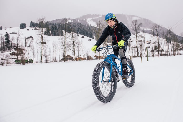 Rider in a blue helmet on a blue fatbike crosses open snow past Gstaad chalets on the Snow Bike Festival prologue.