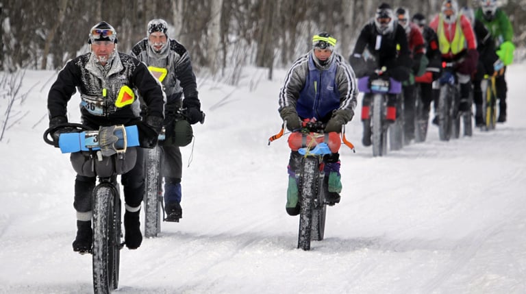 Arrowhead 135 fatbike racers with loaded bikes ride in a line along a snowy trail, faces and jackets covered in frost.