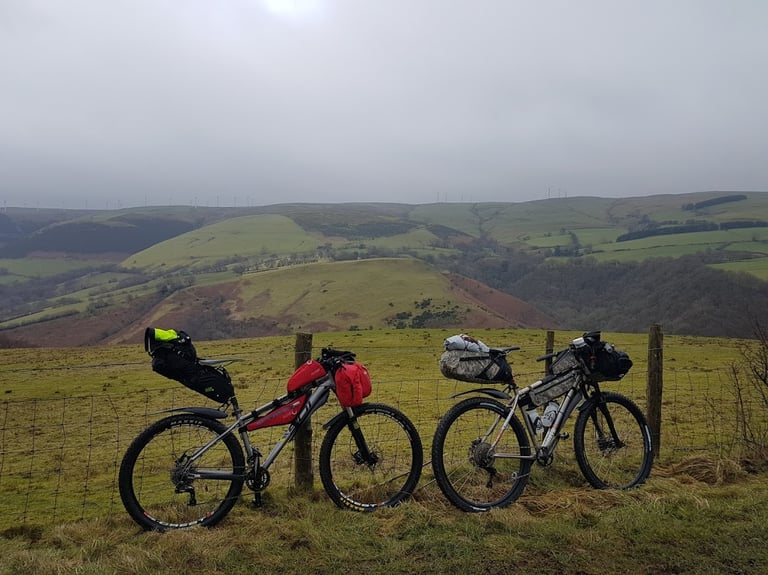 Two loaded bikepacking bikes leaning on a fence above Llanidloes, Powys, with misty hills and wind turbines behind.