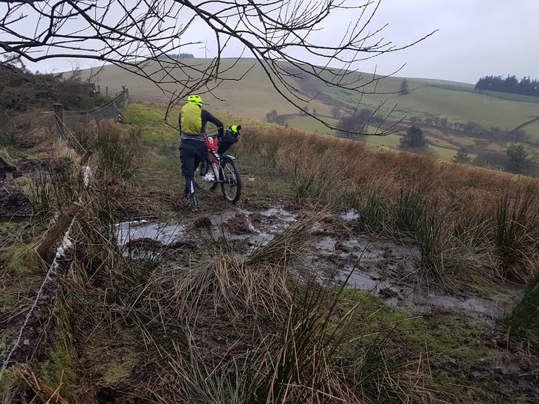 Rider wheels a loaded bike through a flooded, boggy bridleway in Mid-Wales, with wet grass and a wire fence alongside.