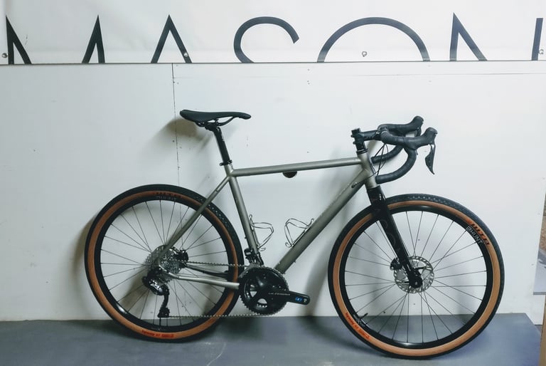 Mason Bokeh Ti titanium gravel bike with tan-wall tyres and Shimano Ultegra cranks, leaning against a white wall.