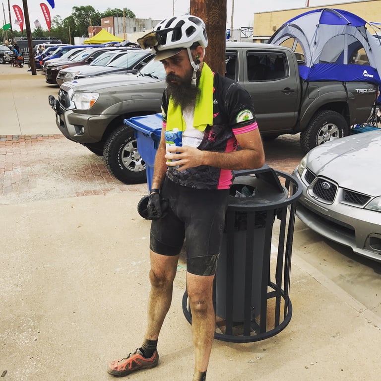 Dust-caked David Markman drinks from a carton beside a bin and parked pickups at a Dirty Kanza 200 checkpoint.