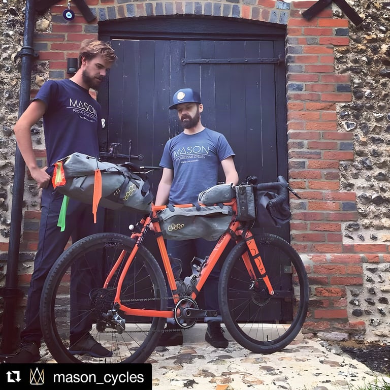 Josh Ibbett and Dom Mason stand behind an orange Bokeh loaded with bikepacking bags at a flint barn doorway.