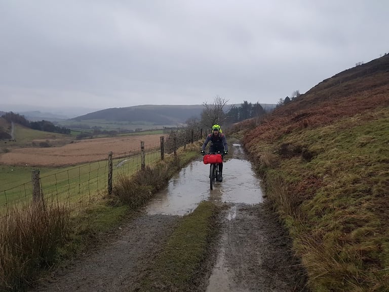 Rider with a red handlebar bag rides through a large puddle on the muddy Pant-y-Dwr climb, Mid-Wales.
