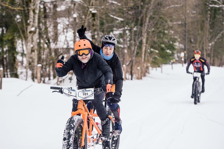 Two riders on an orange fat bike tandem wave and smile on a snowy track at the Snow Bike Festival prologue.