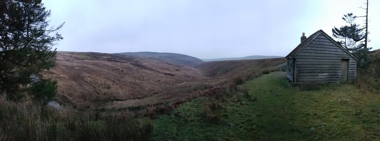 Small wooden bivi hut on a Mid-Wales hillside at dusk, overlooking a wide moorland valley.
