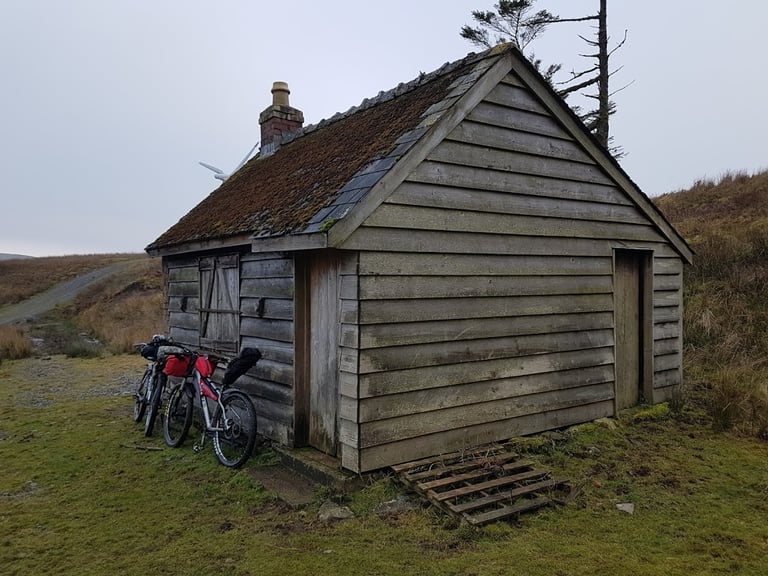 Weathered timber hut with a mossy roof and two loaded bikes leaning on its wall, the Café Racer overnight bivi spot.
