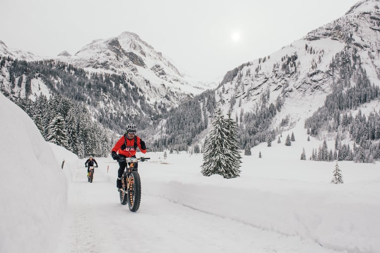 Fatbike rider in a red race top climbs a packed snow road below snowy Alpine peaks on Stage 2.
