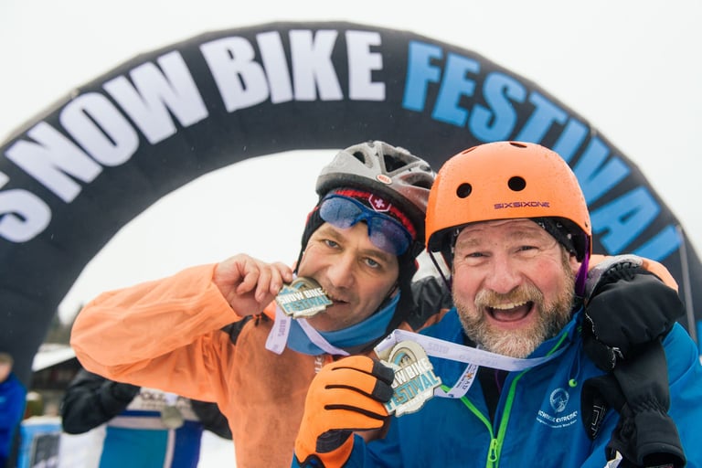 Two finishers bite their medals and grin beneath the Snow Bike Festival inflatable arch in Gstaad.