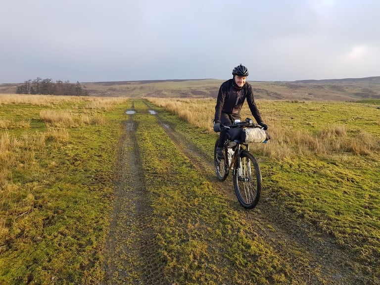 Smiling rider on a loaded bikepacking bike crosses open moorland plateau in low sunlight on the Bearbones Café Racer.