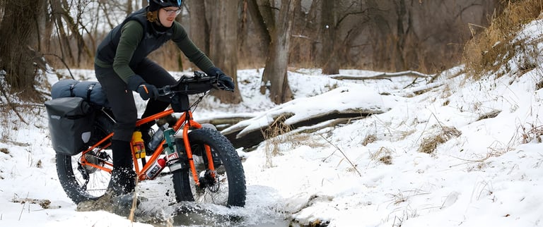 Surly Pugsley V2.0 fatbike with luggage ridden through a snowy stream crossing in woodland.