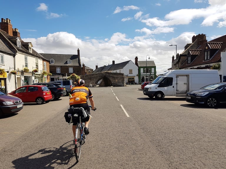 London-Edinburgh-London rider with a saddlebag cycles through a market town square towards a stone bridge.