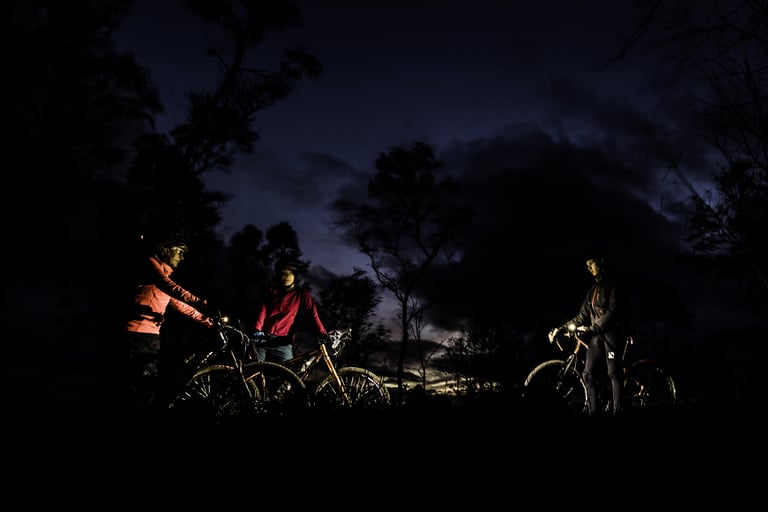 Three riders pause with their bikes under a darkening sky, wrapped up in warm layers, one helmet light glowing.